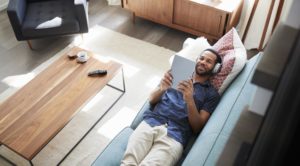 Man Lying On Sofa At Home Wearing Headphones And Watching Movie On Digital Tablet